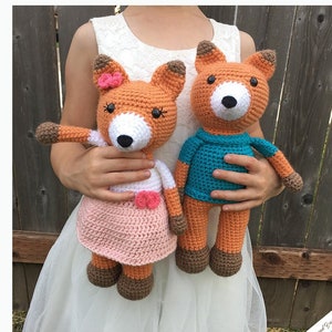 以下が含まれることがあります： ピンクのドレスを着た子とターコイズブルーのシャツを着た子の、2体の編みぐるみキツネ。白いドレスを着た人が抱っこしています。