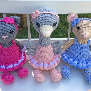 May include: Three crocheted stuffed animals dressed as ballerinas. A gray cat with pink and white tutu, a gray bird with a pink and white tutu, and a tan mouse with a blue and white tutu.