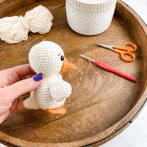 Op de afbeelding: Een witte gehaakte eend met een oranje snavel en poten, garen, een haaknaald en een schaar op een houten dienblad.