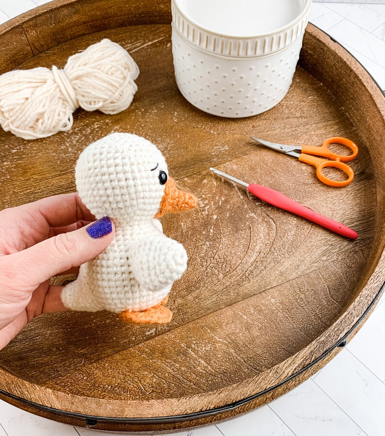 Op de afbeelding: Een witte gehaakte eend met een oranje snavel en poten, garen, een haaknaald en een schaar op een houten dienblad.