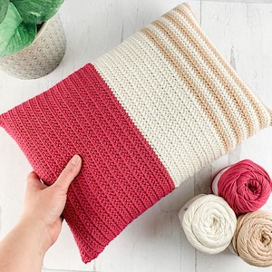 May include: A crocheted pillow with a pink and cream striped design. The pillow is made with a textured stitch and has a square shape.
