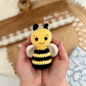 May include: A crocheted bee toy with black and yellow stripes, white wings, and a smiling face. The bee is being held in the palms of two hands.
