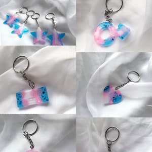 May include: A set of six keychains with different designs. Each keychain features a blue, pink, and white color scheme with glitter accents. The designs include a star, a gender symbol, a heart, a rectangle, and a video game controller.