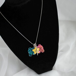 May include: A silver necklace with a pendant featuring two colorful game controllers and a star charm. The controllers are blue and pink with glitter, and the star is yellow. The necklace is displayed on a black mannequin.