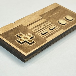 May include: A wooden replica of a Nintendo controller, stained brown. The controller features the classic NES design with a D-pad, select, start, and A and B buttons.