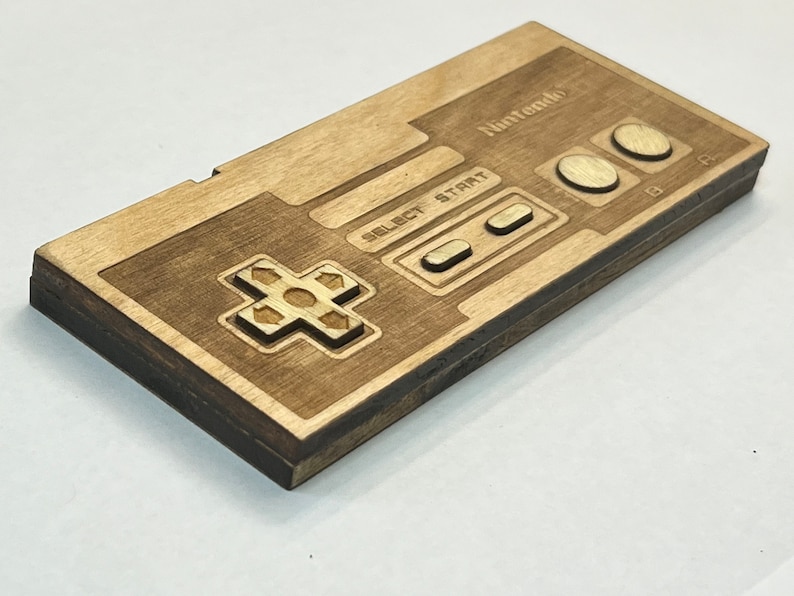 Może przedstawiać: Drewniana replika kontrolera Nintendo z brązowym wykończeniem. Kontroler ma klasyczny design NES z krzyżakiem, przyciskami wyboru, startu oraz A i B.