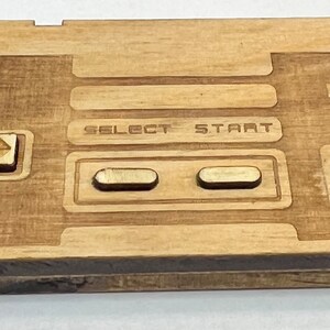 Może przedstawiać: Drewniany kontroler Nintendo z wygrawerowanymi na przodzie napisami "SELECT" i "START". Kontroler wykonany jest z jasnobrązowego drewna i ma naturalne wykończenie.