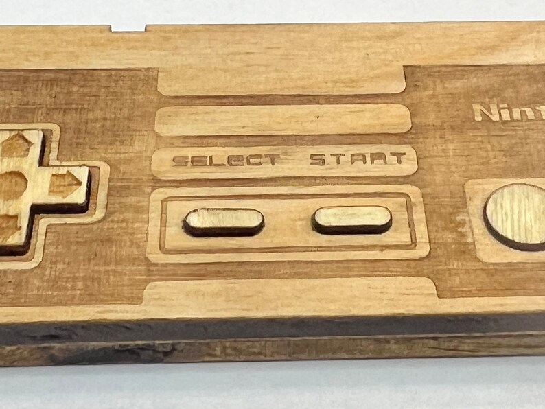 Może przedstawiać: Drewniany kontroler Nintendo z wygrawerowanymi na przodzie napisami "SELECT" i "START". Kontroler wykonany jest z jasnobrązowego drewna i ma naturalne wykończenie.