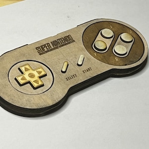 May include: A wooden Super Nintendo controller replica with a brown wood finish and gold accents. The controller features the words "SUPER NINTENDO" on the top, and the buttons "START", "SELECT", "Y", "B", and "A" are labeled.
