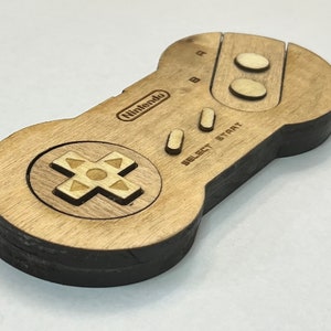 May include: A wooden Nintendo controller with a brown finish. The controller has a D-pad, four buttons, and a "SELECT START" label.