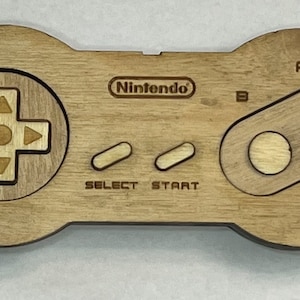 May include: A wooden Nintendo controller with a brown stain. The controller has a D-pad, two buttons labeled "A" and "B", and two buttons labeled "Select" and "Start".