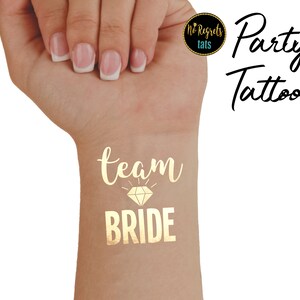 Puede incluir: Tatuaje temporal dorado que dice "Team Bride" con un gráfico de diamante. El tatuaje está en la muñeca de una persona.