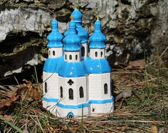May include: A ceramic church figurine with a white body and blue domes. The miniature church has multiple spires topped with gold crosses. The figurine is set in a natural outdoor setting with grass and a tree trunk in the background.