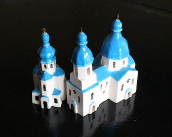 May include: Three miniature white and blue ceramic church buildings with gold crosses on top. The buildings are arranged in a row, with the largest building in the centre.