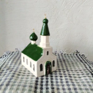 Op de afbeelding: Een klein wit keramieken kerkbeeldje met groene daken en gouden accenten. Het beeldje staat op een geruit tafelkleed.