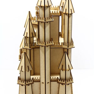 May include: A wooden castle model with multiple towers and intricate details. The castle is made of light brown wood with dark brown lines outlining the details. The castle is standing on a white surface.