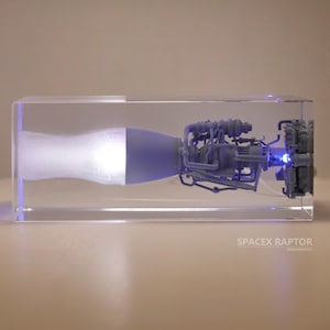 以下が含まれることがあります： 透明なアクリルブロックの中に、スペースXラプターロケットエンジンの詳細なモデルが収められています。エンジンは内側から照らされており、青い光を放っています。ブロックのベースには「SpaceX Raptor」という文字が印刷されています。