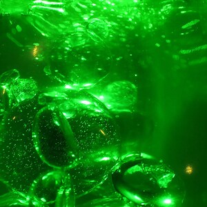 May include: A close-up of a clear glass container filled with a bright green liquid. The liquid is filled with small bubbles and has a shimmering effect.