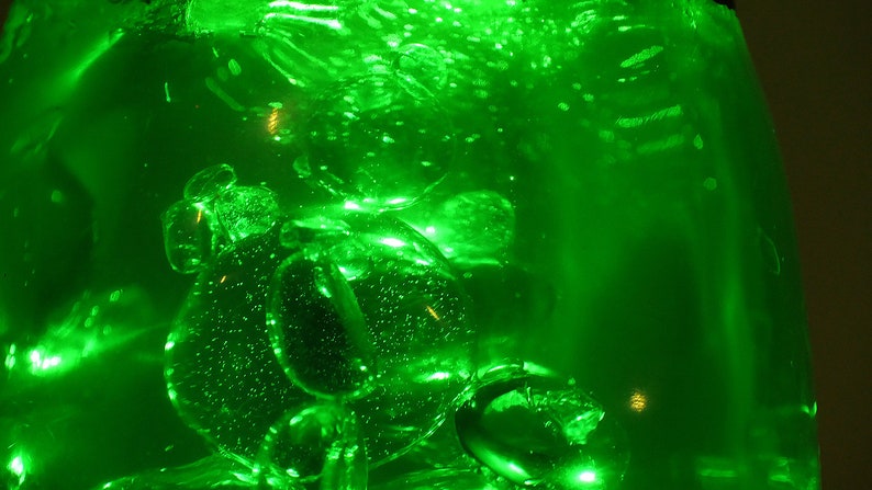 May include: A close-up of a clear glass container filled with a bright green liquid. The liquid is filled with small bubbles and has a shimmering effect.