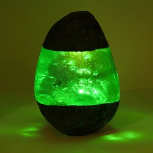 May include: A green resin lamp shaped like an egg with a dark brown base. The lamp is lit and glows green.