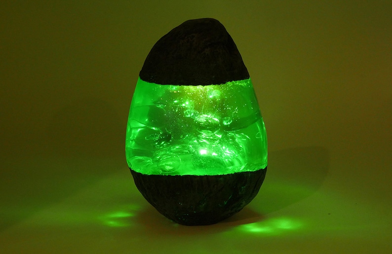 May include: A green resin lamp shaped like an egg with a dark brown base. The lamp is lit and glows green.