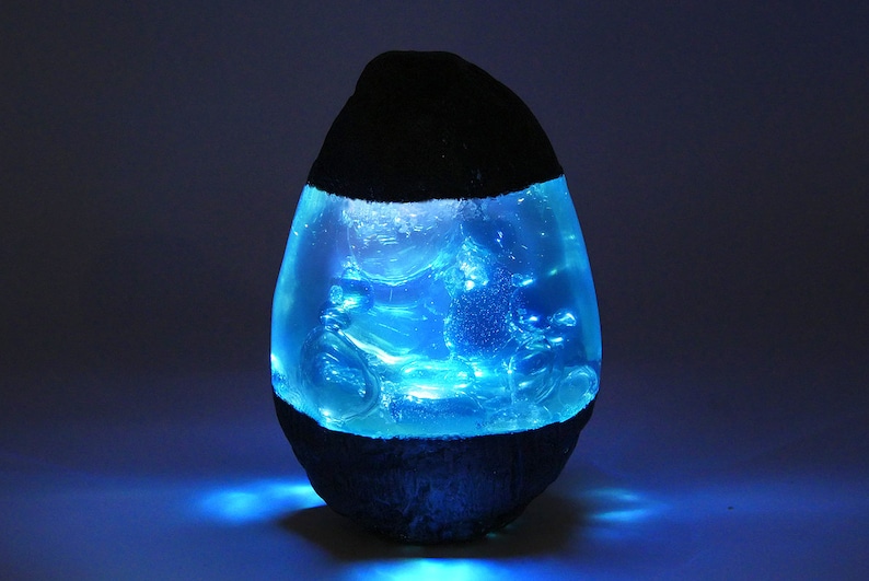 May include: A blue resin egg-shaped lamp with a black base. The lamp is lit and glows blue.