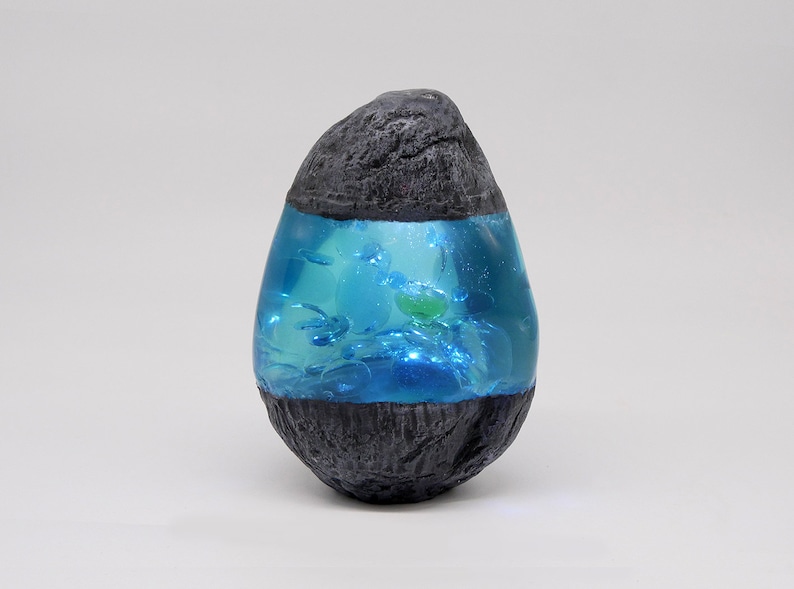 May include: A small, egg-shaped, black rock with a clear blue resin interior containing small, white and green objects.