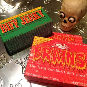 Op de afbeelding: Twee pakketten met nepvoedsel. Het groene pakket zegt "20% Leaner then Doug Jerky! Biff Jerky Human flesh for the Walking dead Net Weight: 8oz" en het rode pakket zegt "Packaged Fresh BRAINS The Treat Zombies Can't resist! Fresh Human Grey Matter, 29979245".
