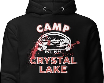Puede incluir: Sudadera con capucha negra con un estampado gráfico blanco de un logotipo de campamento. El logotipo dice "Camp Crystal Lake" con el texto "Est. 1935" debajo. El logotipo está rodeado de salpicaduras de sangre roja.