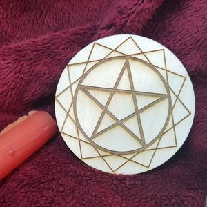 Pode incluir: Um círculo de madeira com um desenho de pentagrama, rodeado por um padrão geométrico. O círculo está sobre uma superfície vermelha e felpuda, ao lado de uma vela branca.