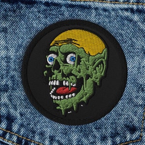 May include: A black and white embroidered patch of a green zombie with yellow hair, with a wide open mouth and large eyes, on a blue denim background.