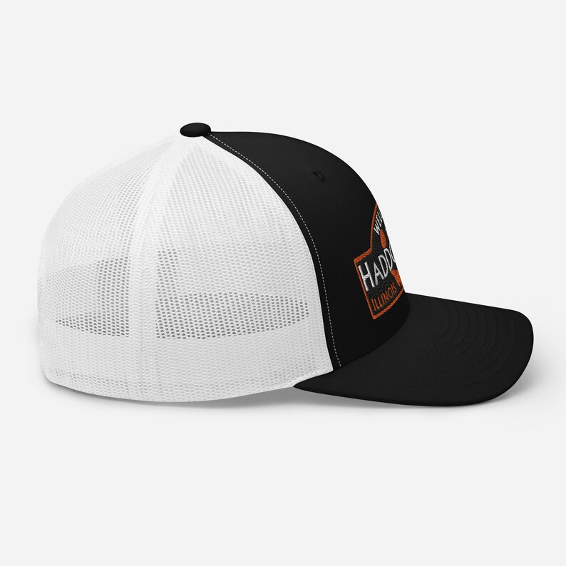 May include: Black and white trucker hat with a white mesh back. The front of the hat features an orange and white logo that says "Westfield Haddorff Illinois".