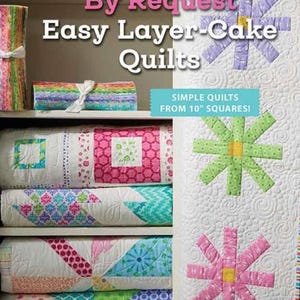 May include: A book cover titled "By Request Easy Layer-Cake Quilts" with stacked quilts and fabric bundles. The book cover features colorful flower designs and the text "Simple Quilts From 10" Squares!" by Barbara Groves & Mary Jacobson.