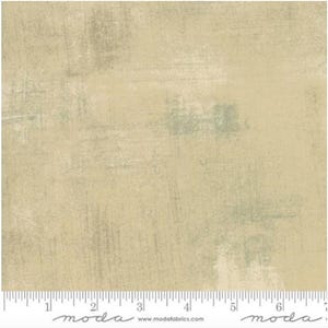 May include: A fabric with a textured, painterly design in shades of beige and muted green. The surface has visible brushstroke-like patterns, creating a distressed, artistic look. The bottom edge features a ruler with inch measurements and the word "moda".