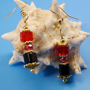 May include: A pair of dangle earrings with gold-colored hooks. Each earring features a red square bead, a cluster of iridescent beads, and a black square bead, all accented with gold-colored filigree and beads. The earrings are displayed against a seashell.