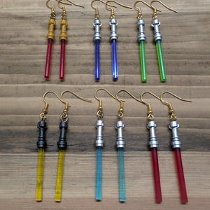 May include: A set of 10 lightsaber earrings. The earrings are made of gold, silver, and black metal with coloured plastic lightsabers. The lightsabers are red, blue, green, yellow, and turquoise.