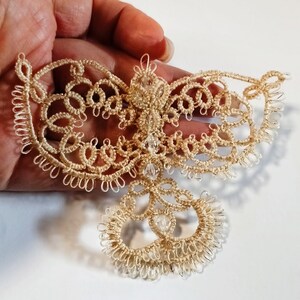 Pu&ograve; includere: Un delicato ornamento in pizzo beige fatto a mano con un design simmetrico. L'ornamento presenta intricati motivi ad anello ed &egrave; adornato con piccole perle trasparenti. Il pizzo &egrave; tenuto in mano su sfondo bianco.