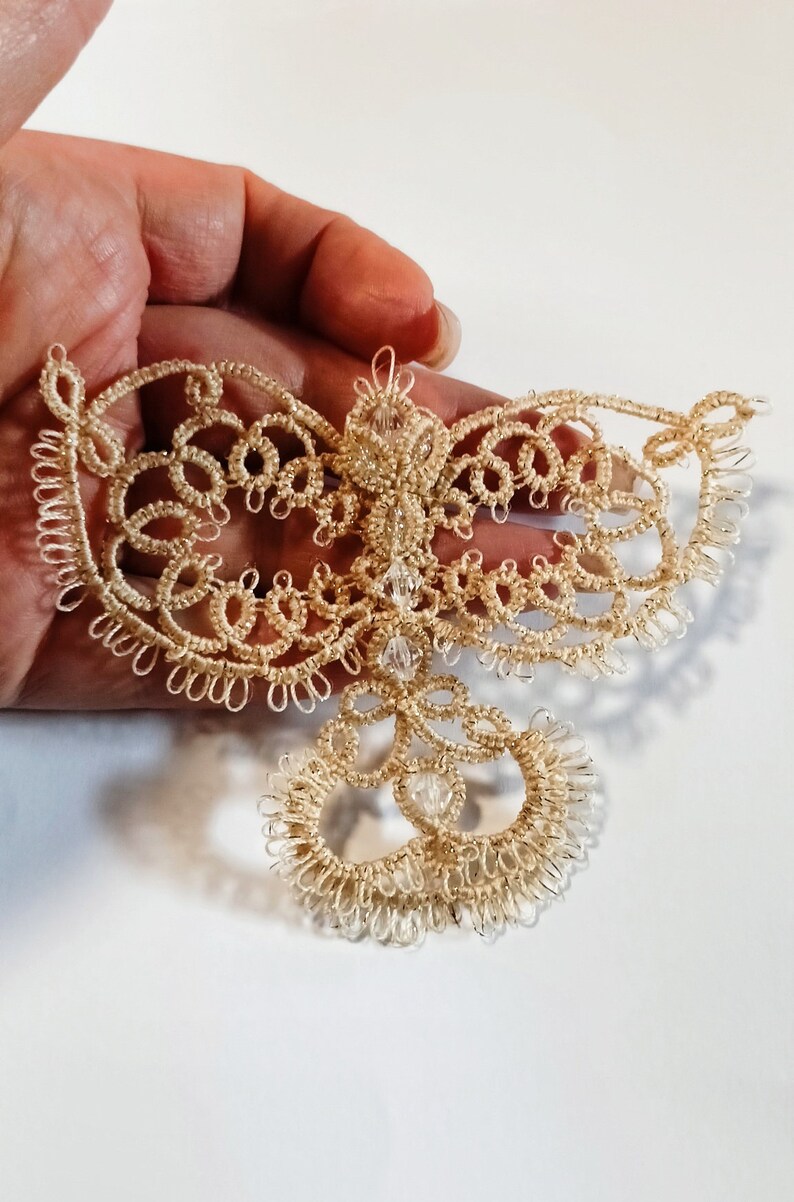Pu&ograve; includere: Un delicato ornamento in pizzo beige fatto a mano con un design simmetrico. L'ornamento presenta intricati motivi ad anello ed &egrave; adornato con piccole perle trasparenti. Il pizzo &egrave; tenuto in mano su sfondo bianco.