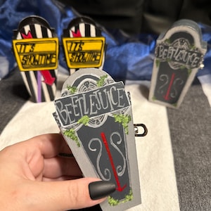 May include: Three black and white striped coffin-shaped ornaments. Two have yellow signs that read "It's Showtime" and the third has a gray sign that reads "Beetlejuice".