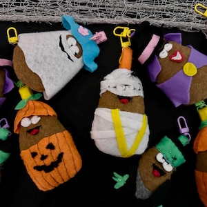 May include: A collection of felt potato-shaped keychains featuring various Halloween costumes including a mummy wrapped in white fabric, a pumpkin with a jack-o'-lantern face, a witch with a purple cape and hat, and a ghost with a blue hat.