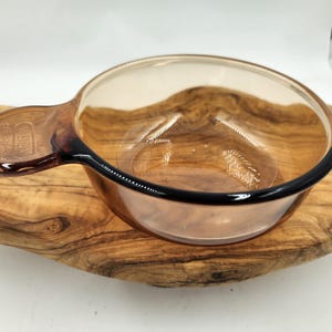 May include: A brown glass baking dish with a handle. The dish is sitting on a wooden cutting board.