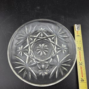May include: Clear glass plate with a starburst pattern. The plate is round and has a diameter of approximately 6 inches. A yellow and black measuring tape is next to the plate.