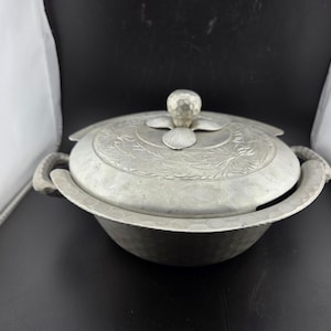 May include: A silver-colored metal serving dish with a lid. The dish has a round bowl shape with two handles and a decorative floral pattern on the lid. The lid has a knob on top. The dish appears to be vintage.