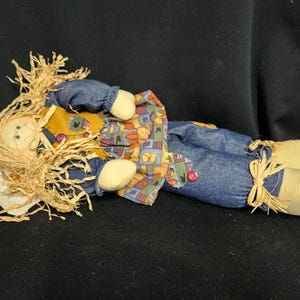 May include: A handmade rag doll with a straw-like hair, wearing a blue denim outfit with a patchwork skirt and brown boots. The doll is lying on its side with its head resting on a white pillow.