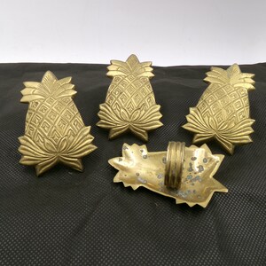 May include: Set of four gold pineapple napkin rings. The rings are made of metal and have a detailed pineapple design. One ring is shown from the back, revealing a simple ring.