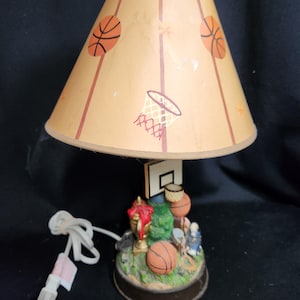 May include: A table lamp with a beige lampshade featuring basketballs and a basketball hoop. The base of the lamp is a 3D sculpture of a basketball scene with a trophy, basketballs, and a player.