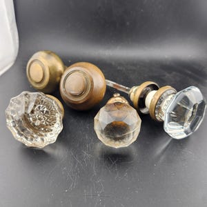 May include: A collection of vintage door knobs in various styles. The knobs include brass, wood, and clear glass with faceted and floral designs. The knobs are arranged on a dark surface, showcasing their unique shapes and textures.