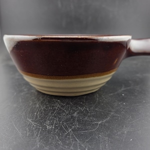 May include: A brown ceramic soup bowl with a handle. The bowl has a dark brown interior and rim, with a tan band and base. The handle is also brown. The bowl is designed for serving soups and stews.
