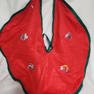 May include: Red felt Christmas tree skirt with a green trim. The skirt features embroidered images of Santa Claus, reindeer, and other Christmas characters.