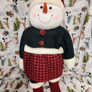 May include: A plush snowman doll wearing a green jacket, red and white plaid skirt, and a red hat with a green holly sprig. The snowman has a white face with black eyes, a carrot nose, and a red smile.